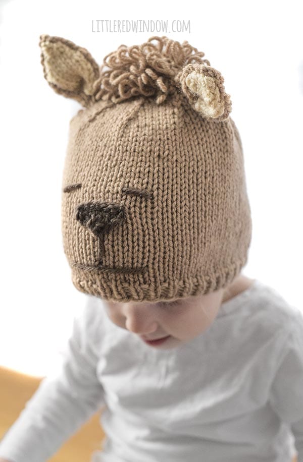 little girl looking down and to the left and wearing tan alpaca hat with brown nose and curly hair on top