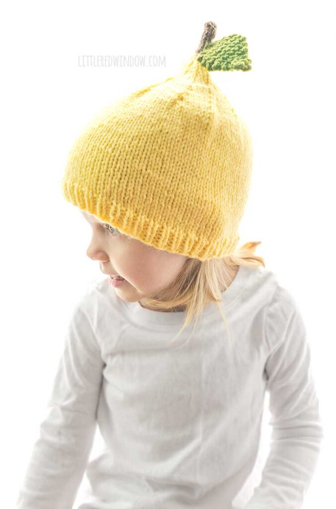 closeup of little girl wearing yellow knit lemon hat with stem and leaf and looking off to the left in front of a white background