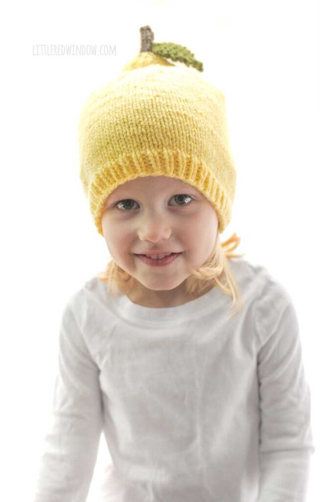 little girl in white shirt wearing yellow lemon knit hat with stem and leaf and smiling at the camera in front of a white background