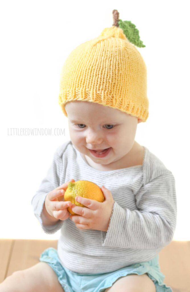 Smiling Baby sitting and holding a lemon and wearing a yellow knit lemon hat