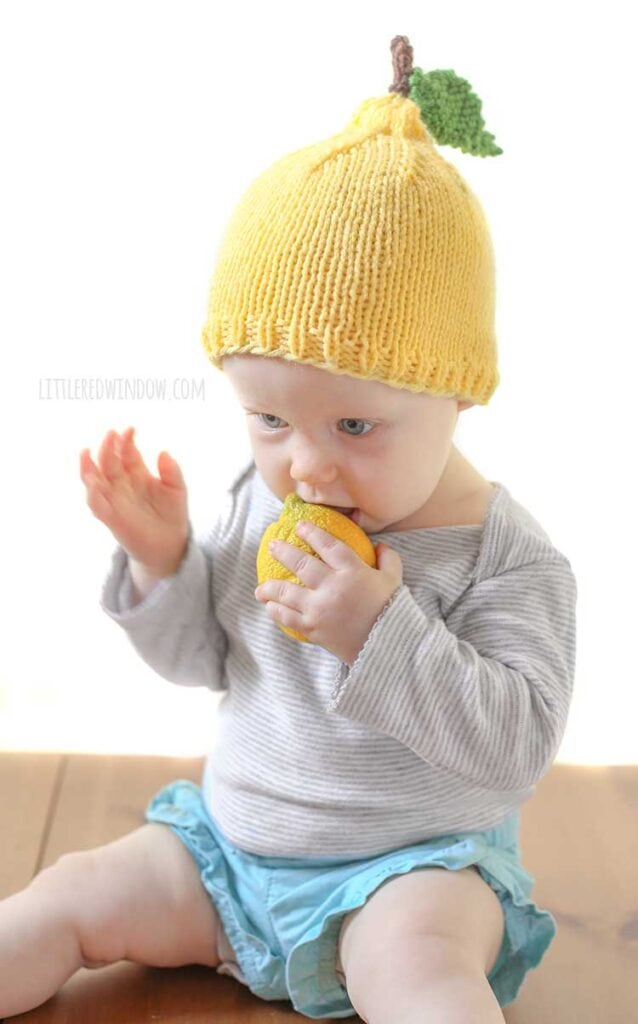 baby putting mouth on whole lemon and wearing a yellow knit lemon hat