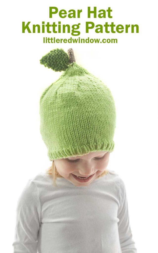 girl in white shirt wearing a light green knit hat that looks like a pear with a stem and leaf on top sitting front of a white background and looking down at her lap