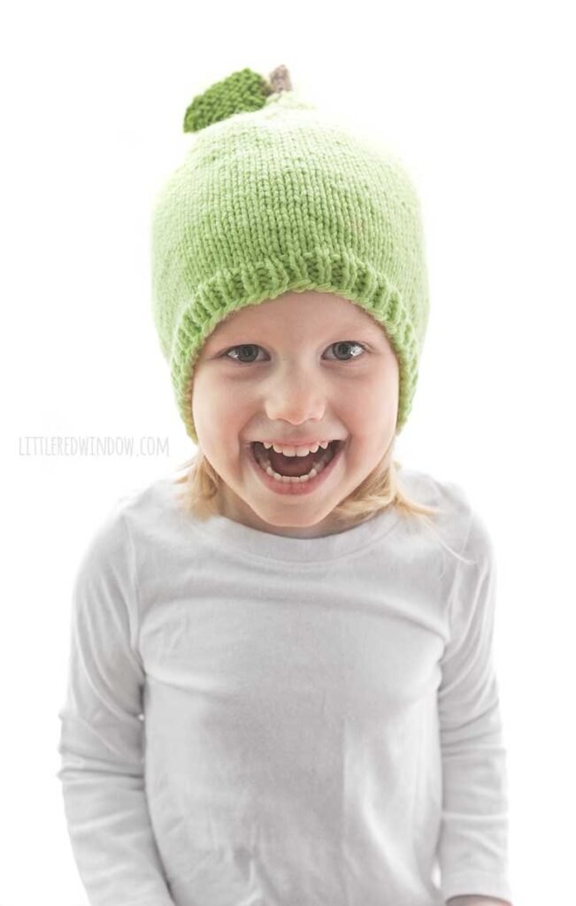 girl in white shirt wearing a light green knit hat that looks like a pear with a stem and leaf on top sitting front of a white background and laughing at the camera