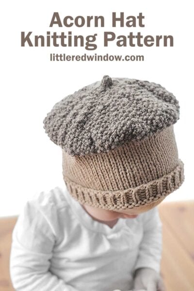 A baby wearing a knitted acorn hat and white shirt looks downward. The hat, with its textured top resembling an acorn cap, captures a rustic charm. Above, text reads: "Acorn Hat Knitting Pattern" and "littleredwindow.com.
