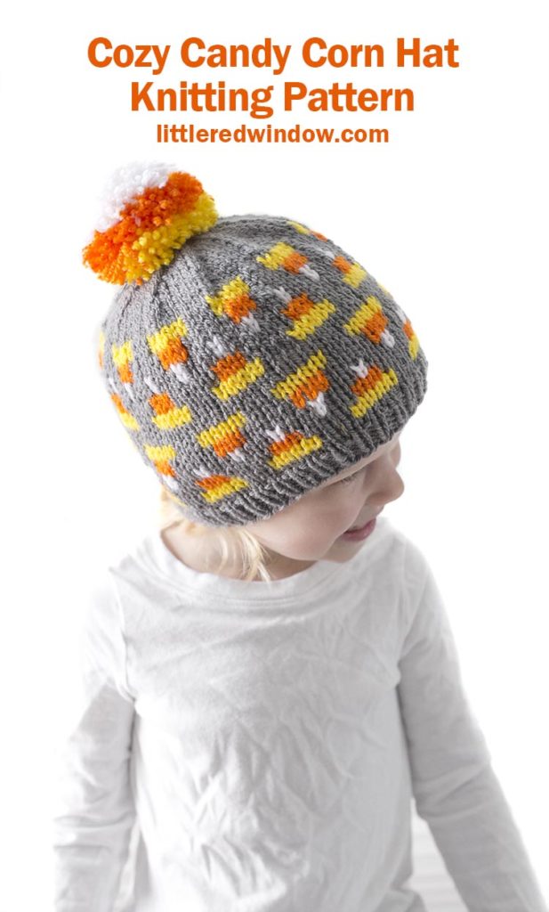 little girl in white shirt looking to the right and wearing gray knit hat with candy corn pattern on it and candy corn colored pom pom on top