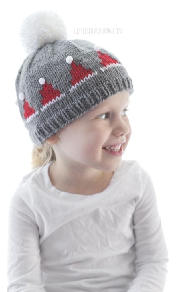 little girl wearing gray knit hat with small red santa hats on it and looking off to the right