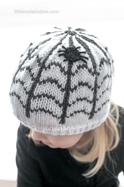 A child wearing a white knit hat featuring a black spiderweb design and a small spider on top.