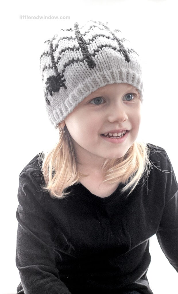 A young child wearing a hand-knitted beanie with a spider web and spider pattern.