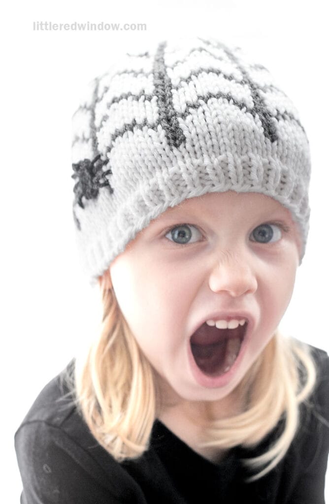 A child wearing a light grey hand-knitted beanie with a dark grey spiderweb pattern and a small spider on the side, looking surprised with their mouth open.