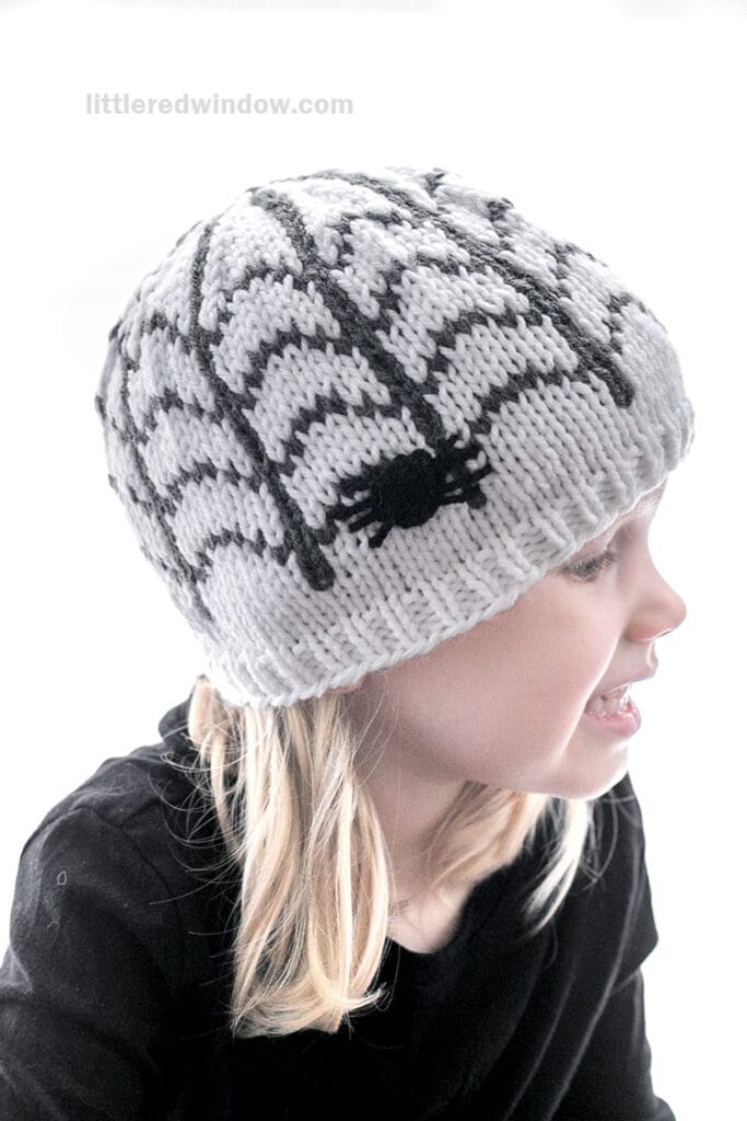 A child wearing a knitted beanie featuring a spider web and spider pattern looking to the right.