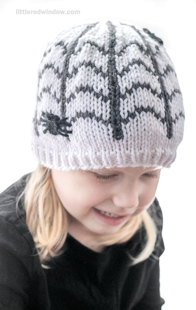 A young child wearing a white knit hat featuring a black spiderweb pattern and a small spider.