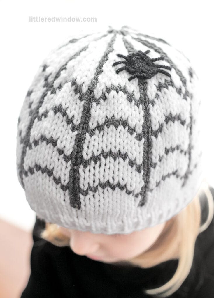 A child wearing a white knit hat featuring a black spider web pattern and a small black spider applique.