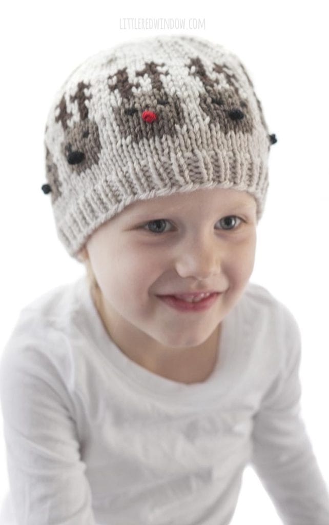 little girl smiling and wearing tan knit hat with team of reindeer heads around the center