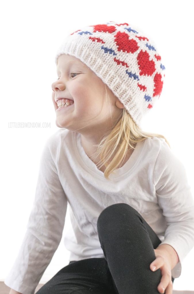 little girl sitting cross legged and wearing a white knit hat with red hearts and blue and red airmail stripes