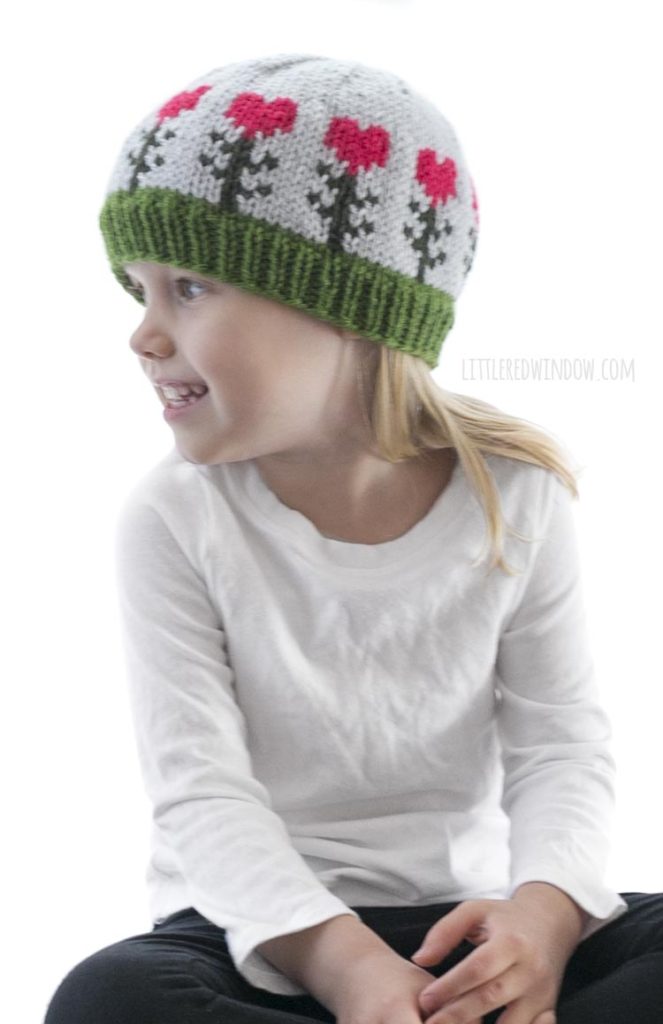 little girl in white shirt wearing light blue knit hat with pink heart shaped flowers on it looking off to the left