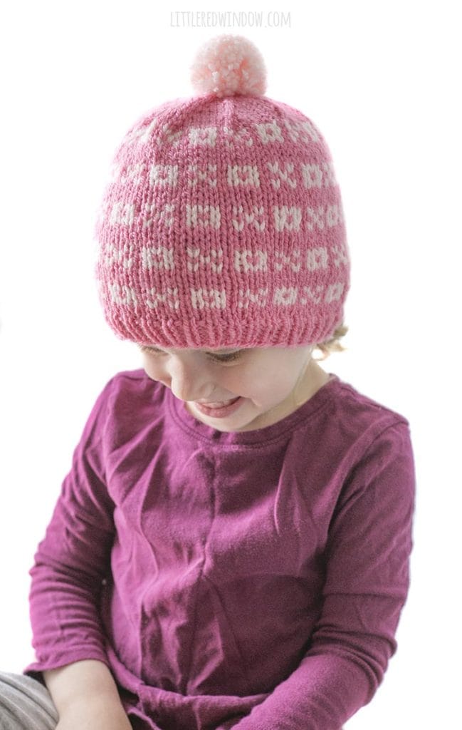 little girl in purple shirt wearing a light pink knit hat with alternating pattern of xs and os