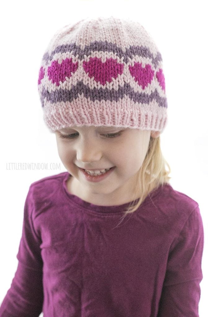 little girl in purple shirt wearing a pink knit hat with dark pink hearts and purple scallops