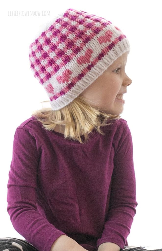 little girl in purple shirt wearing hat with pink hearts and pink gingham plaid looking off to the right