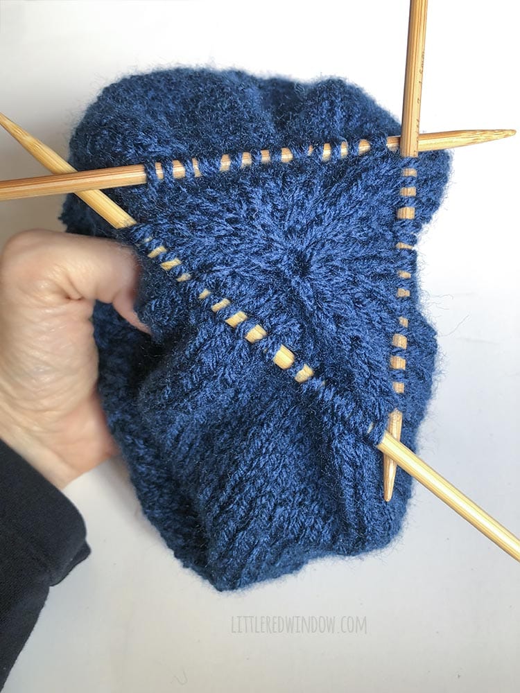 hand holding a blue knit hat with a round of picked up stitches on double pointed knitting needles near the crown