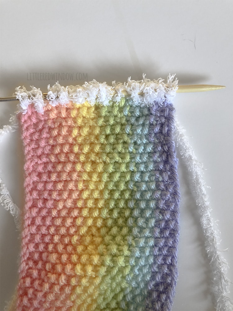 rainbow colored scarf with a knitting needle picking up new live stitches with white yarn along one short end