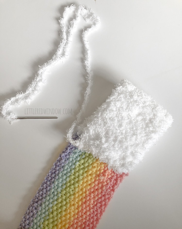 rainbow scarf with white cloud section folded over on itself to make a loop with yarn end threaded on a yarn needle