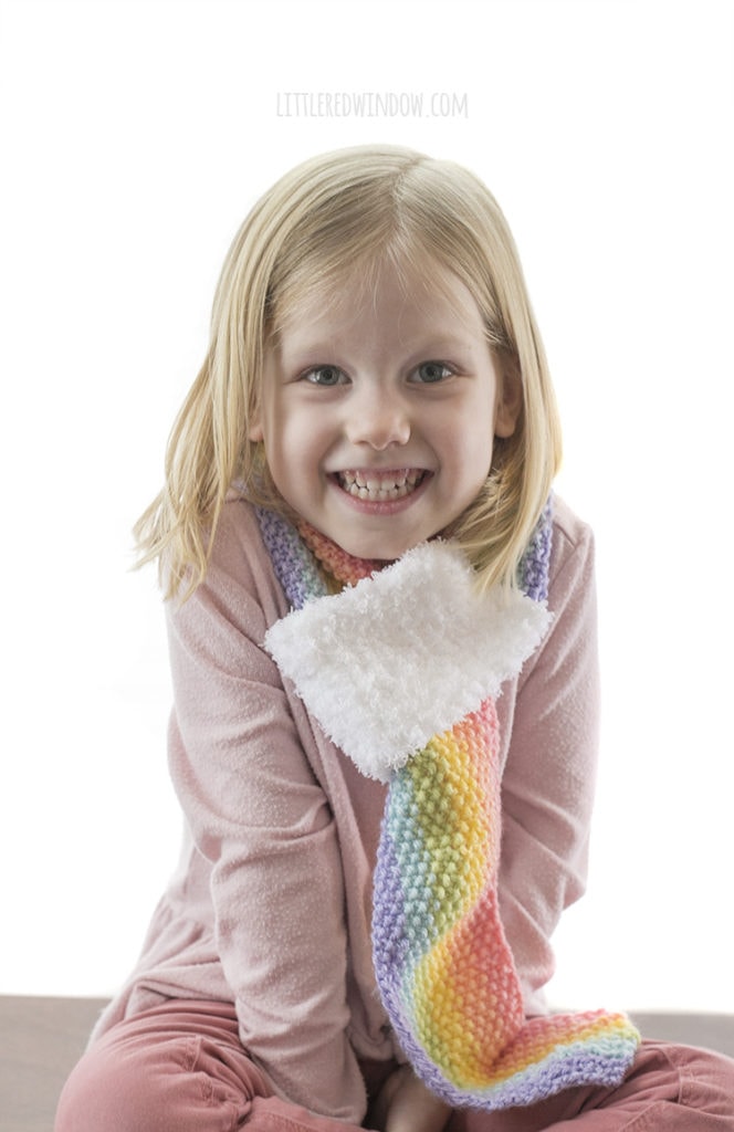 little girl in pink shirt smiling at the camer and wearing a rainbow striped knit scarf in front of a white background