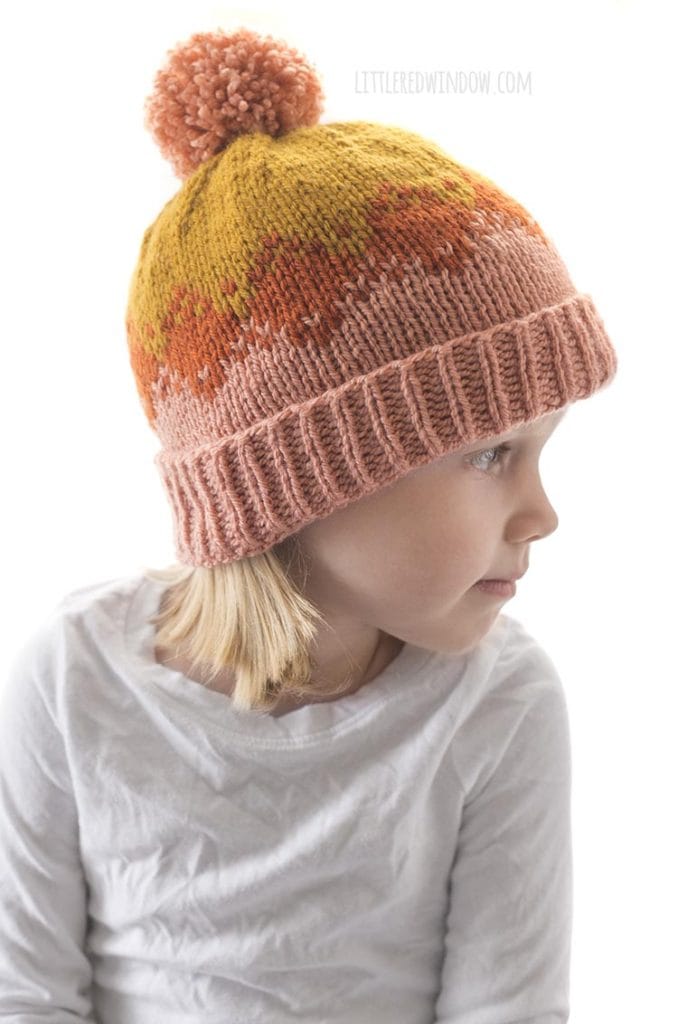little blond girl in white shirt wearing pink rust and mustard colored autumn fade hat knitting pattern and looking off to the right