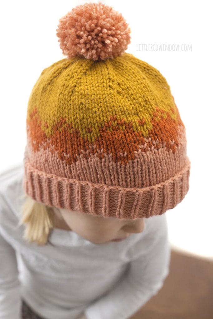 top view of little girl in white shirt wearing pink orange and mustard autumn fade hat knitting pattern and looking down