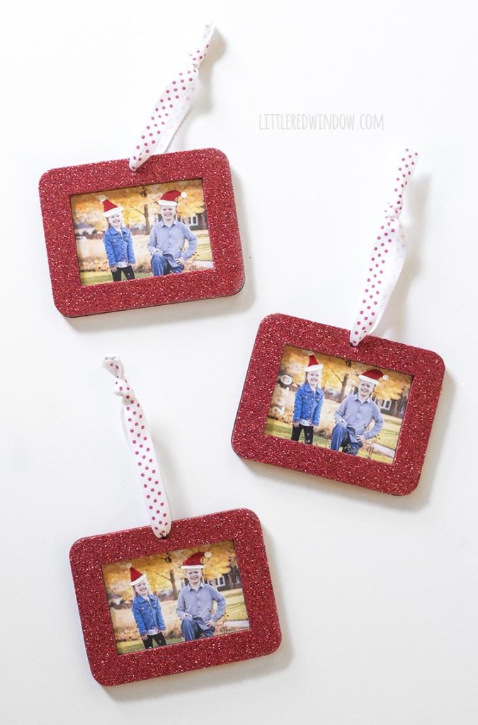 three red rectangular glitter photo ornaments with a photo of two kids with painted on santa hats inside on a white background
