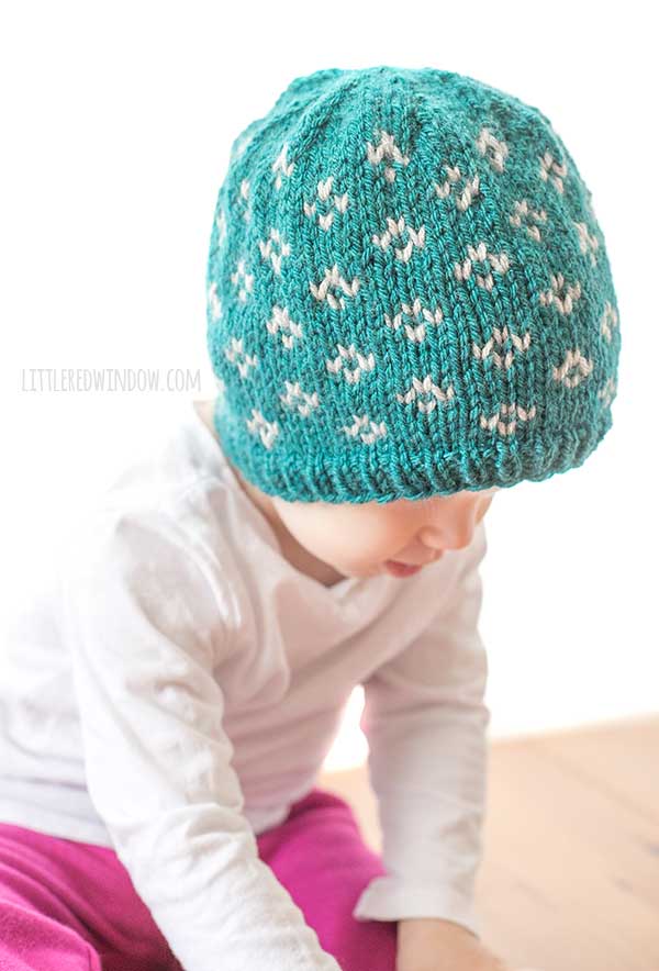 little baby in white shirt leaning over to crawl and wearing a teal knit hat with light gray little flowers diamond pattern on it