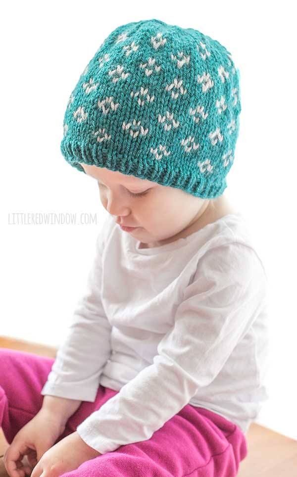 baby in white shirt and hot pink pants sitting cross legged and wearing a teal knit little flowers hat with light gray flower diamond shapes on it