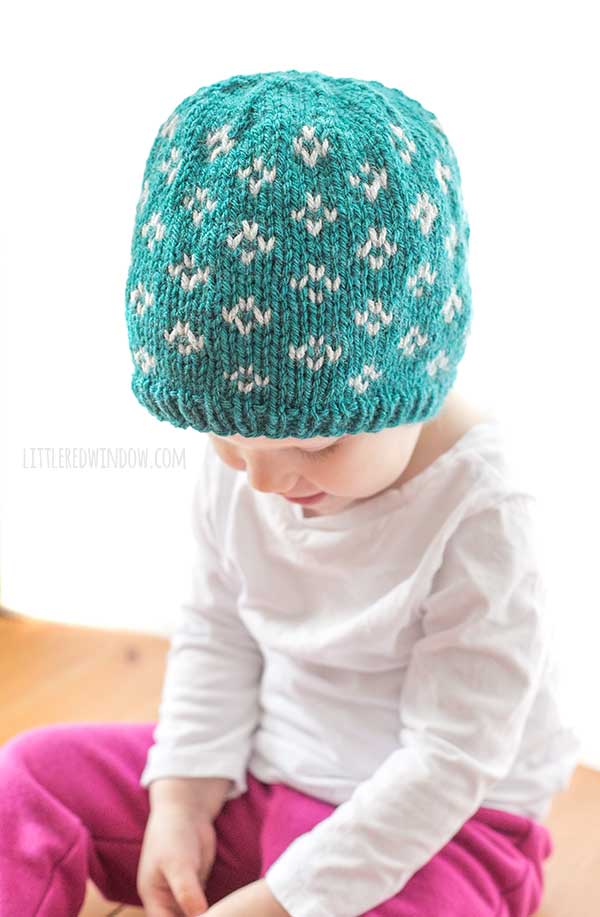 baby in white shirt looking down at their lap and wearing a teal knit hat with light gray little flower diamond pattern on it