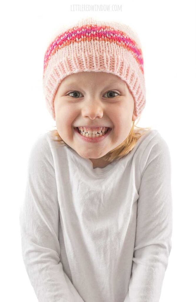 girl in white shirt smiling and wearing diagonal stripe fiery fade hat in light pink orange magenta and purple fade