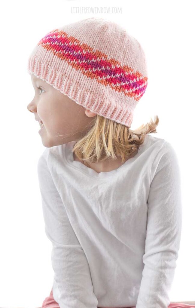 girl in white shirt looking to the left and wearing diagonal stripe fiery fade hat in light pink orange magenta and purple fade