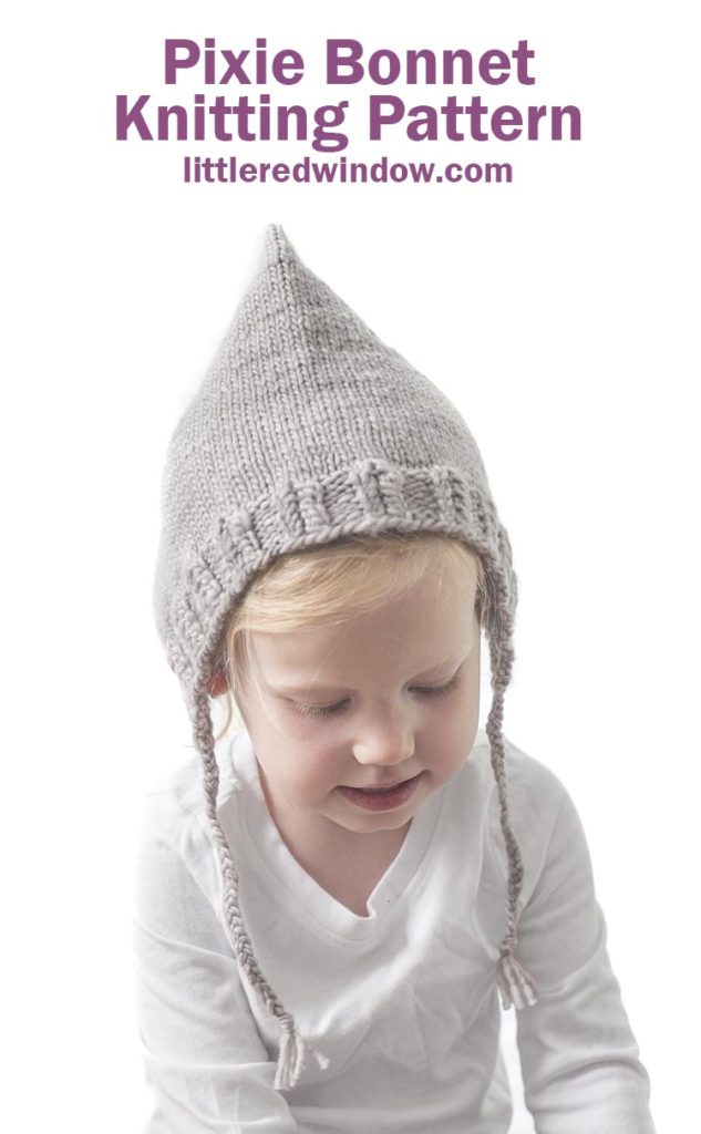 small toddler in white shirt wearing tan pointed knit pixie bonnet with braided ties and looking down