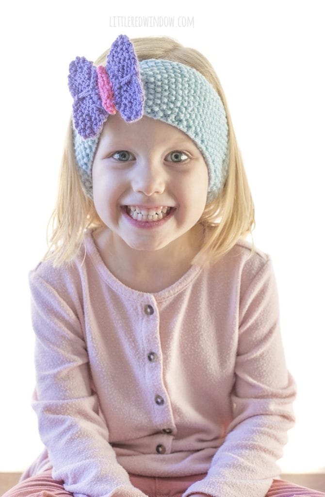 little girl in pink shirt wearing blue knit headband with purple butterfly on the front looking right at the camera