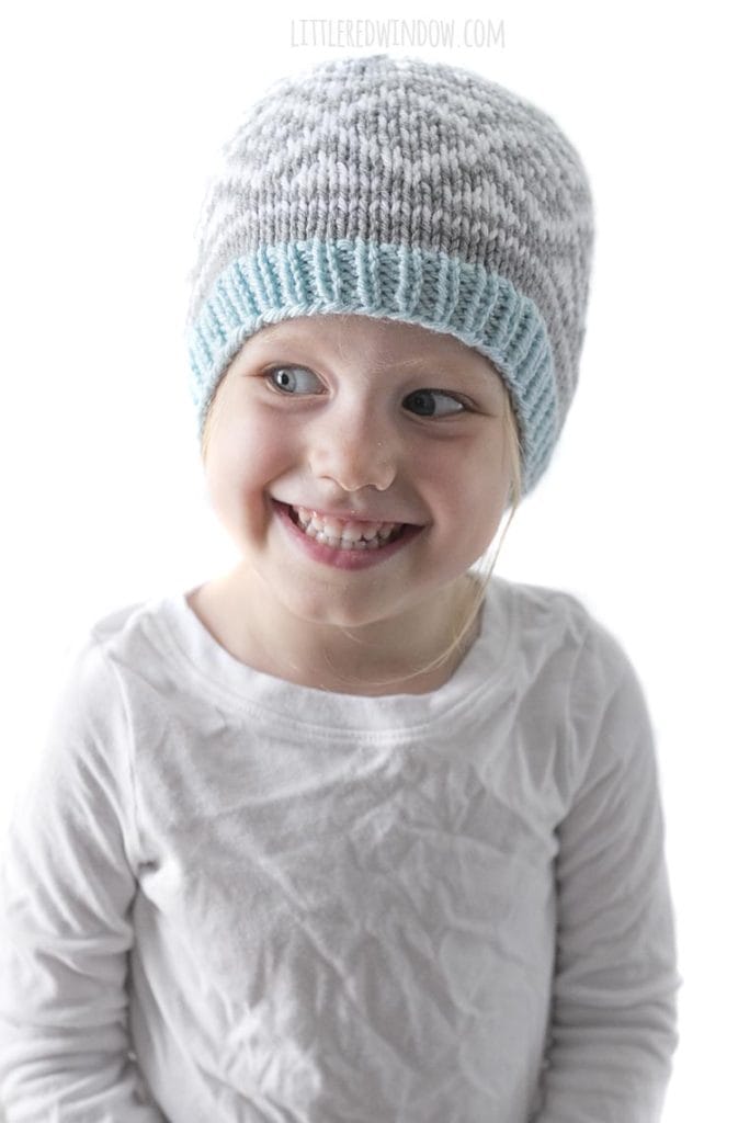 smiling girl in white shirt wearing gray and white diamond patterned knit hat and looking to the left