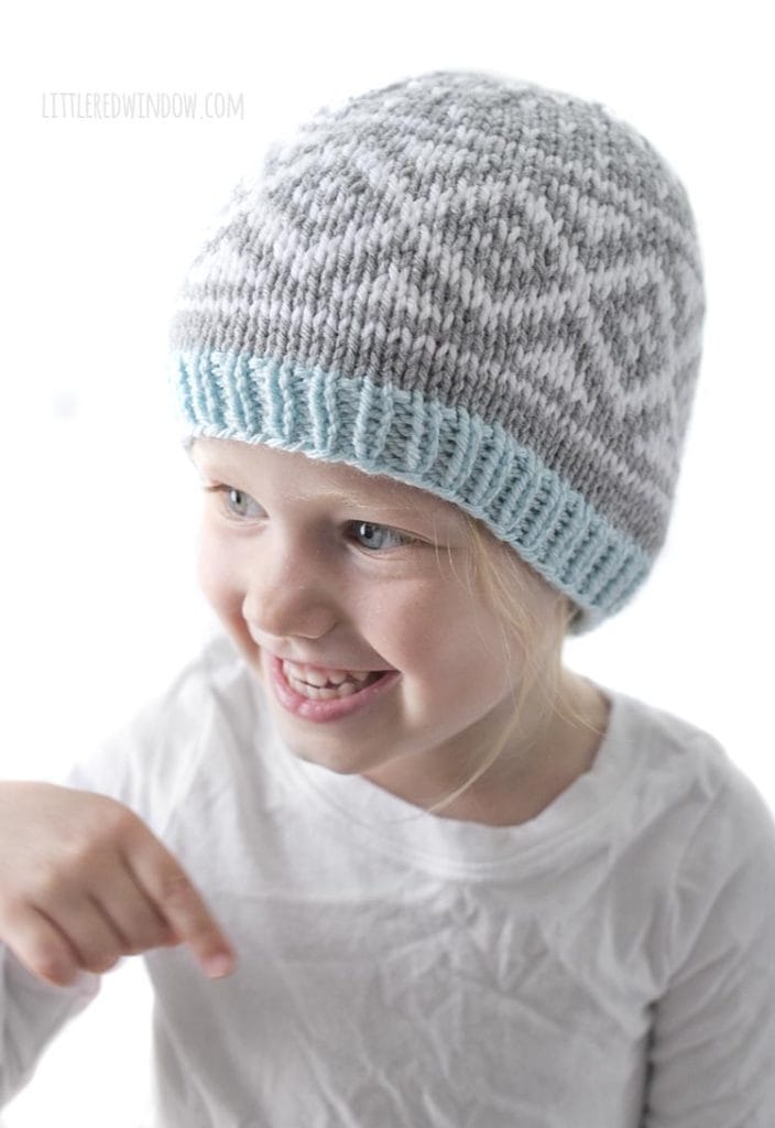 smiling girl from above wearing a white shirt and gray and white diamond patterned knit hat with blue brim