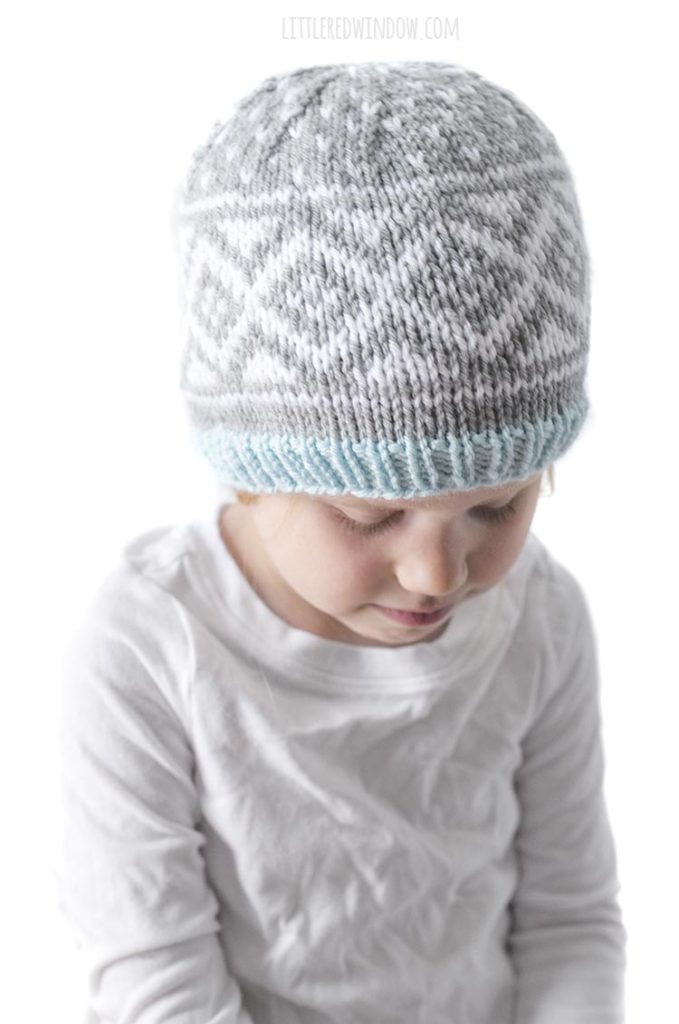little girl in white shirt looking down at her lap and wearing knit hat with gray and white diamond pattern and blue ribbed brim
