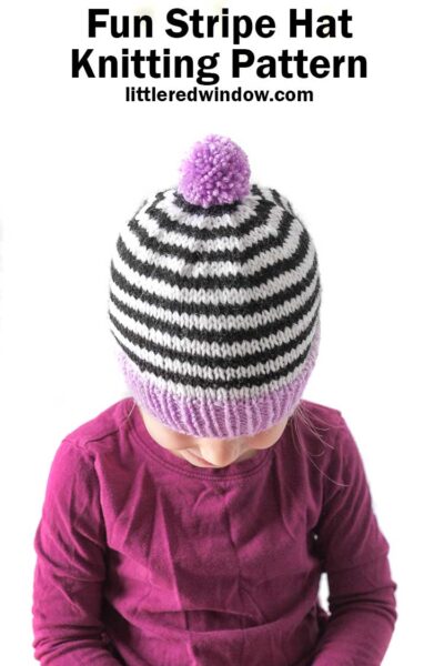 little girl in magenta shirt wearing black and white striped knit hat with purple brim and pom pom looking down at her lap