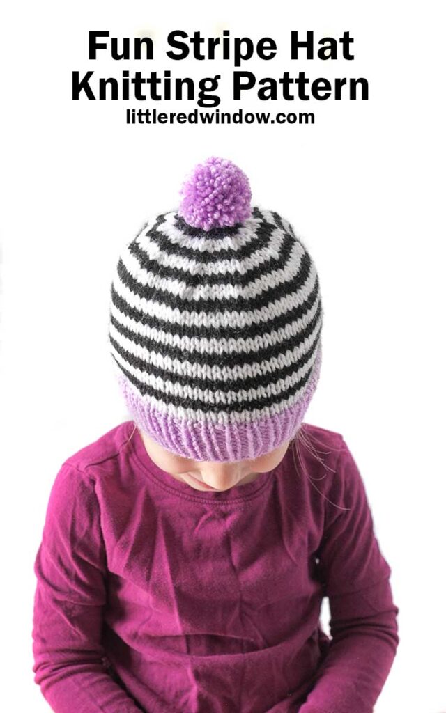 little girl in magenta shirt wearing black and white striped knit hat with purple brim and pom pom looking down at her lap