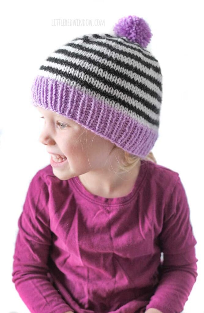 little girl in magenta shirt wearing black and white striped knit hat with purple brim and pom pom looking off to the left