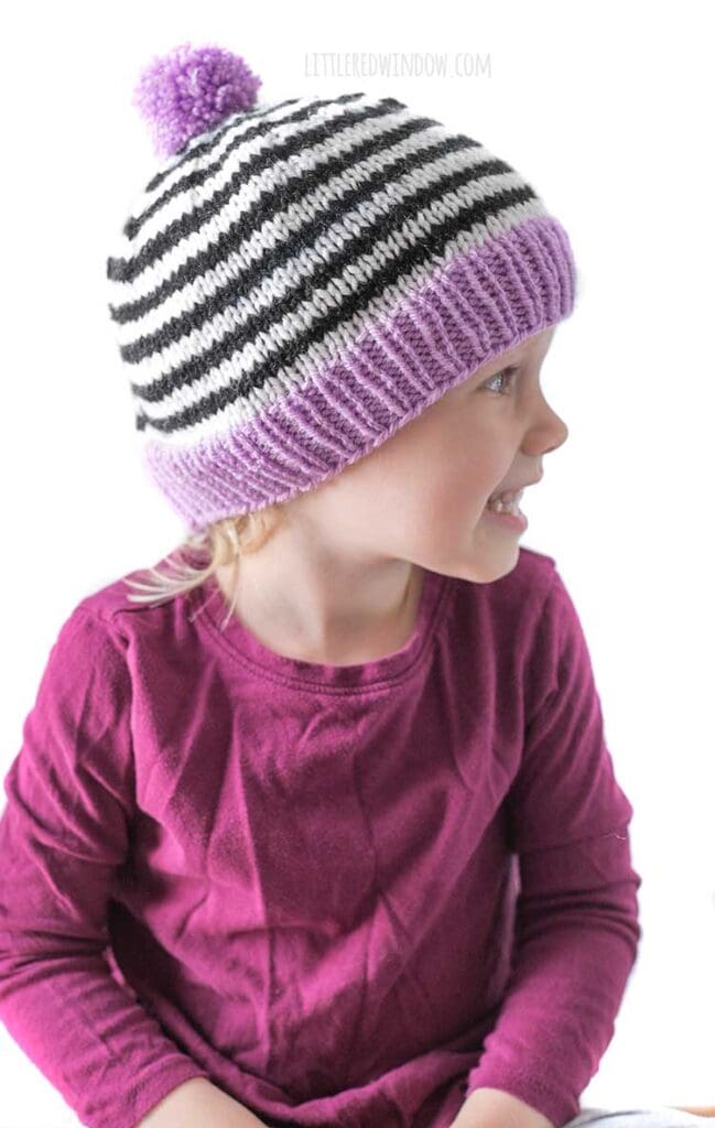 little girl in magenta shirt wearing black and white striped knit hat with purple brim and pom pom looking off to the right