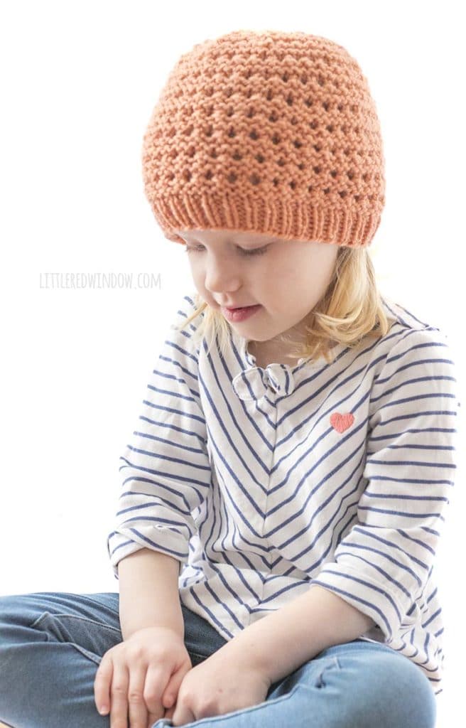 little girl in white and navy shirt and coral colored knit lace grid hat looking down at her hands in her lap