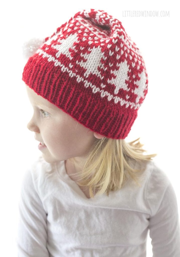 little girl in red and white fair isle stocking cap with tree motifs around the middle looking off to the left