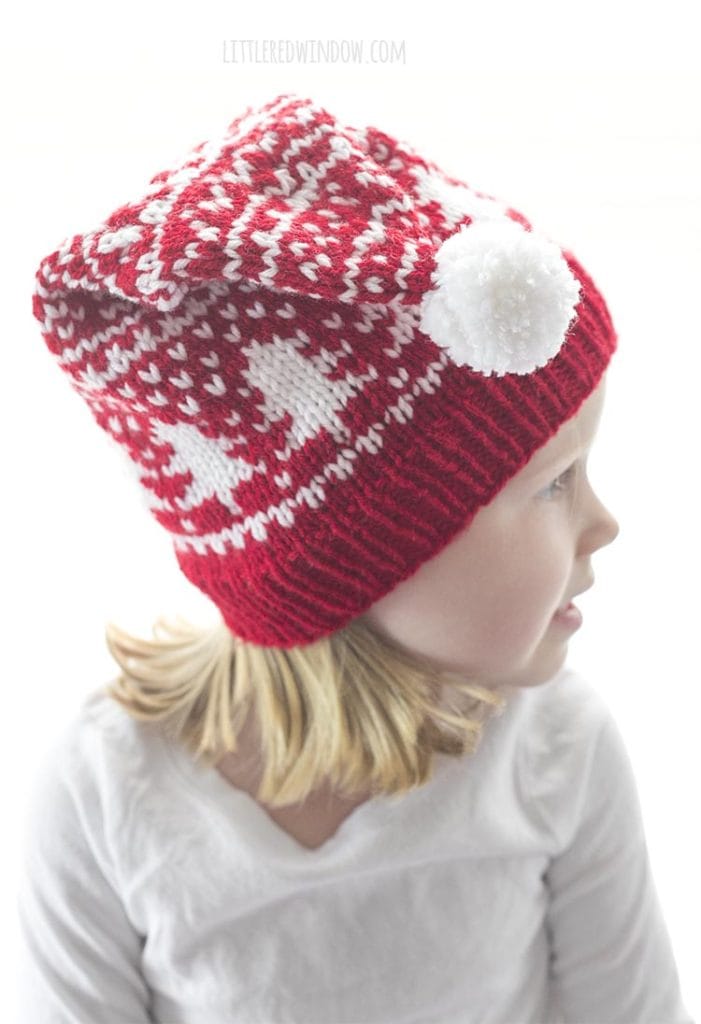 little girl in red and white fair isle stocking cap with tree motifs around the middle and a white pom pom on the top looking off to the left