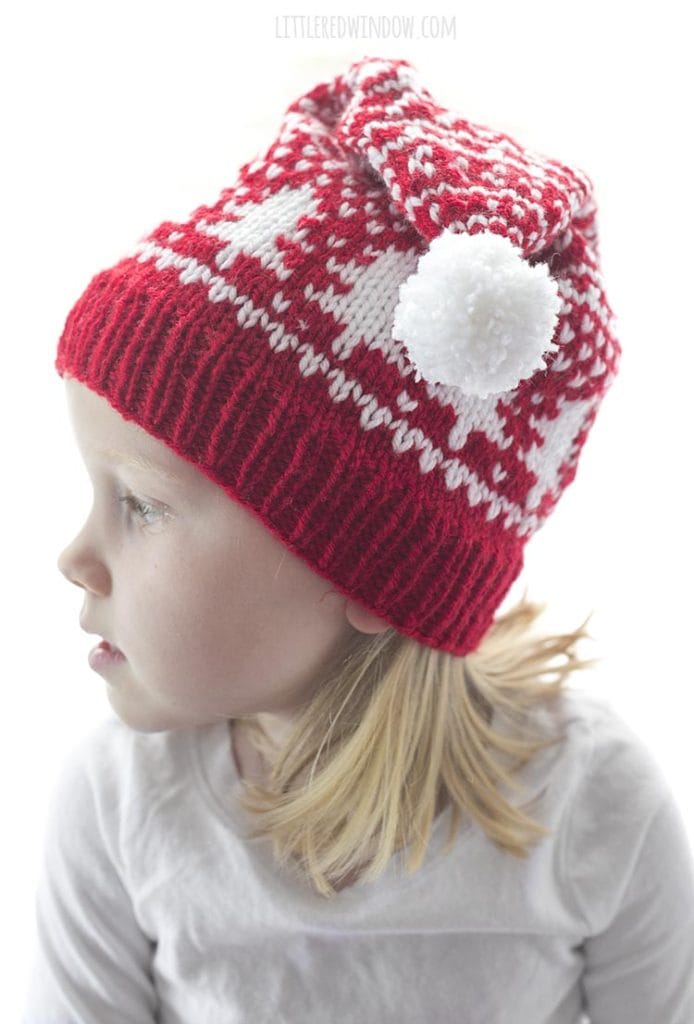 little girl in red and white fair isle stocking cap with tree motifs around the middle and a white pom pom flopped over to the side looking off to the left