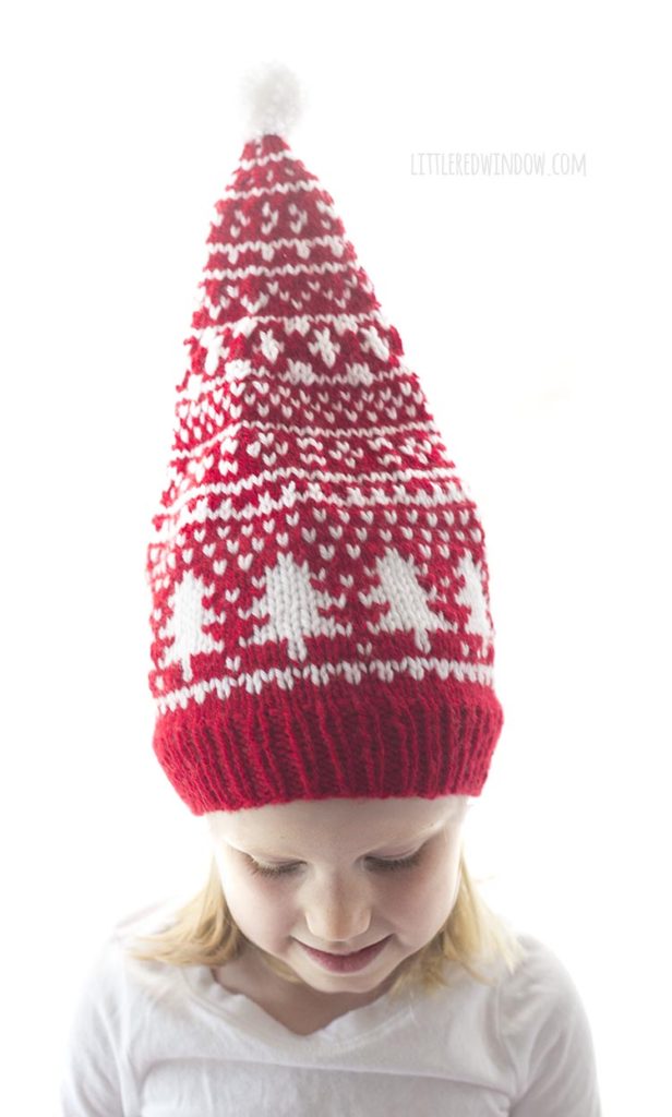 little girl in white shirt looking down wearing red and white fair isle stocking cap with stocking cap sticking straight up in the air to show all of the fair isle designs and the white pom pom on top