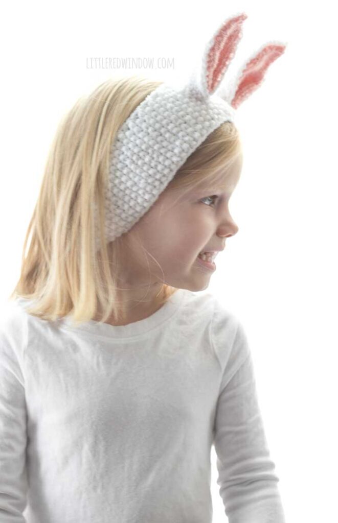 little blond girl in white shirt wearing seed stitch knit white headband with white and pink bunny ears on top looking off to her right