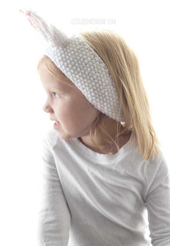 little blond girl in white shirt wearing seed stitch knit white headband with white and pink bunny ears on top looking off to her left